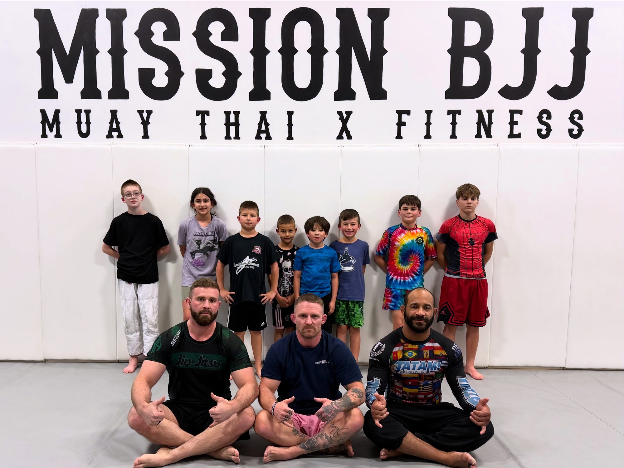 Kids martial arts class posing with their coaches beneath the Mission BJJ wall logo in Mission, BC